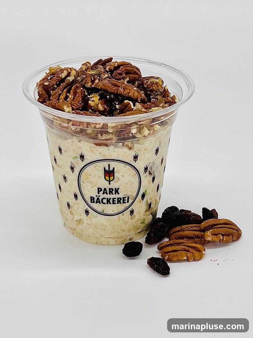 A clear plastic cup filled with overnight oats, topped with glazed pecans. The cup has a label with the logo "PARK BÄCKEREI" and a German flag emblem. A small pile of raisins and pecans is scattered next to the cup.