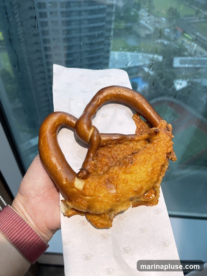 A hand wearing a striped watch holds a pretzel that appears to be filled with a golden-brown, crispy substance. The pretzel is resting on white paper with a repeating logo. In the background, through a window, a blurred cityscape with tall buildings is visible.