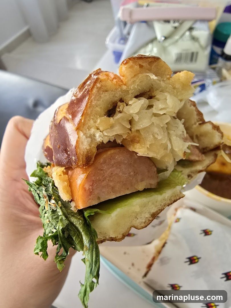 A hand holds a sandwich with a pretzel bun, sausage, lettuce, and sauerkraut. In the background, there are blurred items and packaging with a logo.