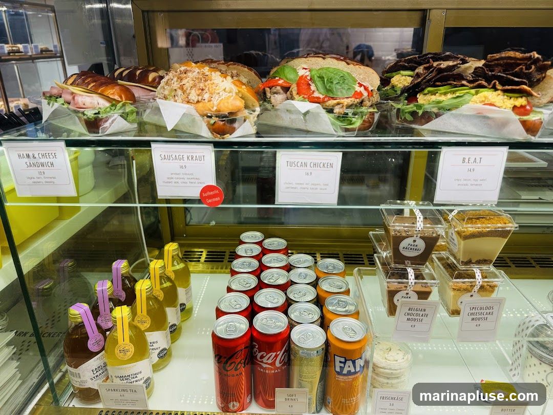 A refrigerated display case filled with various food items including sandwiches, desserts, drinks, and canned beverages. The sandwiches are labeled with their names and prices. Desserts include chocolate mousse and cheesecake mousse. Several bottles of sparkling tea and cans of soda are also visible.
