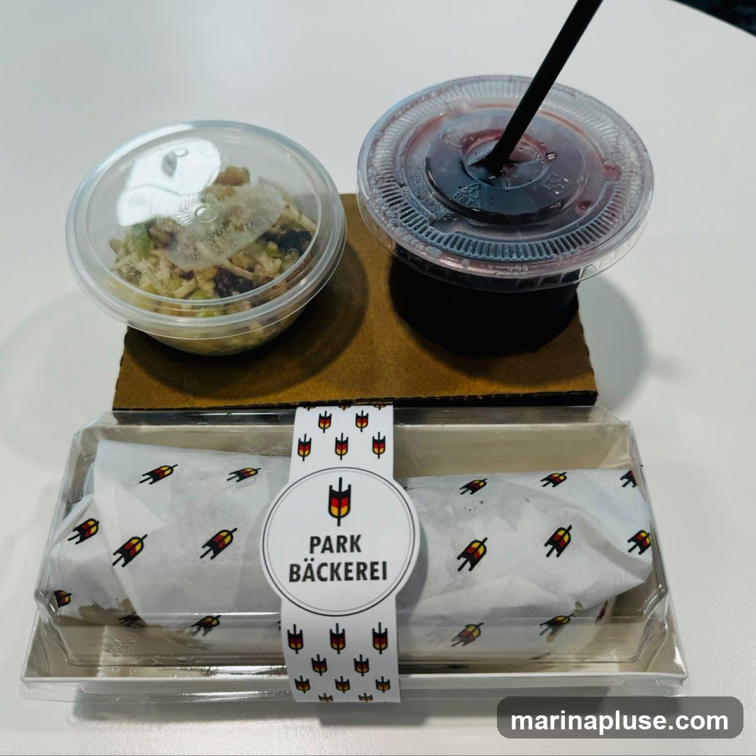 A takeaway meal consisting of a sandwich wrapped in branded paper, a small container of salad, and a cup of dark-colored beverage with a straw. The sandwich is in a clear plastic container, and the salad and drink are in clear plastic cups with lids. The paper wrapping the sandwich has a repeating pattern of small figures and the logo "PARK BÄCKEREI" is visible on a circular sticker.