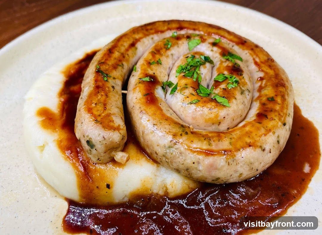 A close-up shot of a coiled sausage served on a bed of mashed potatoes, drizzled with a rich brown sauce and garnished with chopped parsley. The sausage appears to be grilled or pan-fried, with visible herbs and spices. The mashed potatoes are smooth and creamy, and the sauce has a glossy sheen. The dish is presented on a light-colored, textured plate.