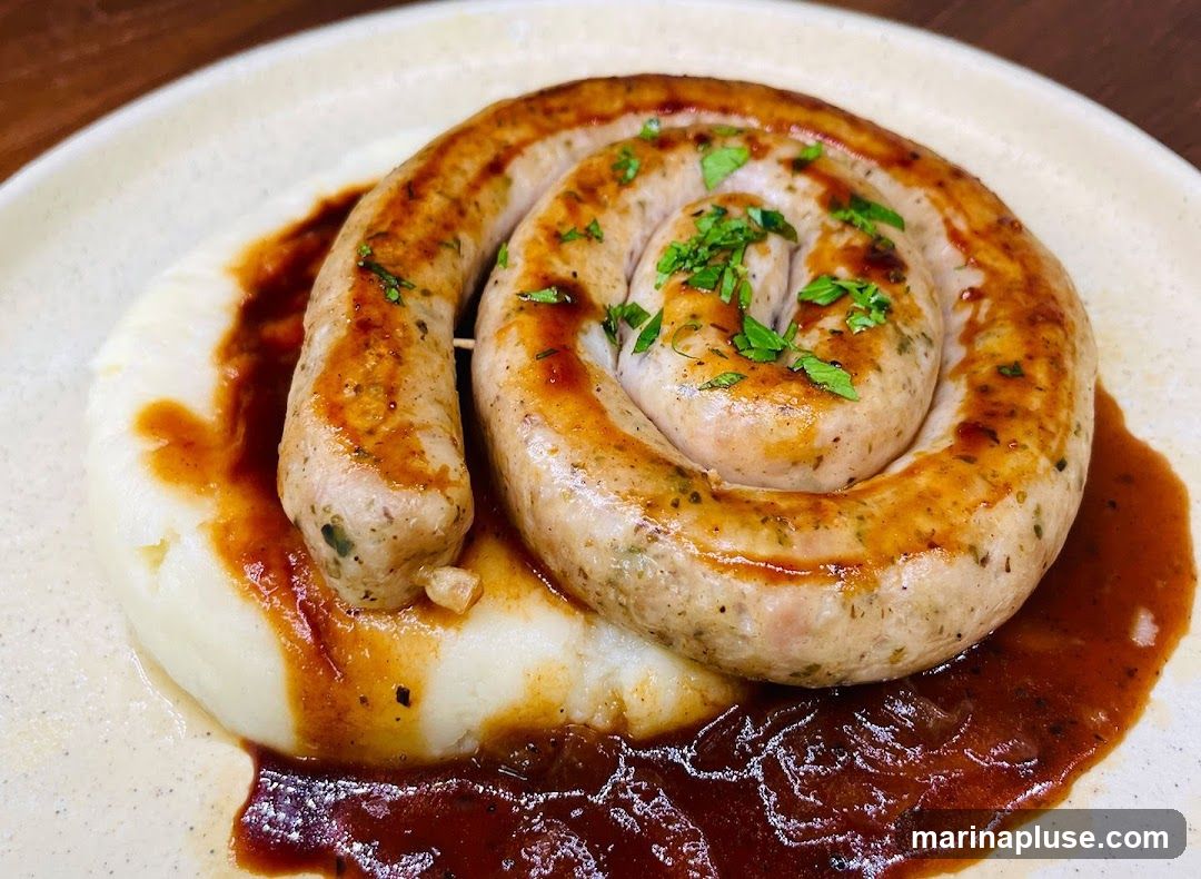 A close-up shot of a coiled sausage served on a bed of mashed potatoes, drizzled with a rich brown sauce and garnished with chopped parsley. The sausage appears to be grilled or pan-fried, with visible herbs and spices. The mashed potatoes are smooth and creamy, and the sauce has a glossy sheen. The dish is presented on a light-colored, textured plate.
