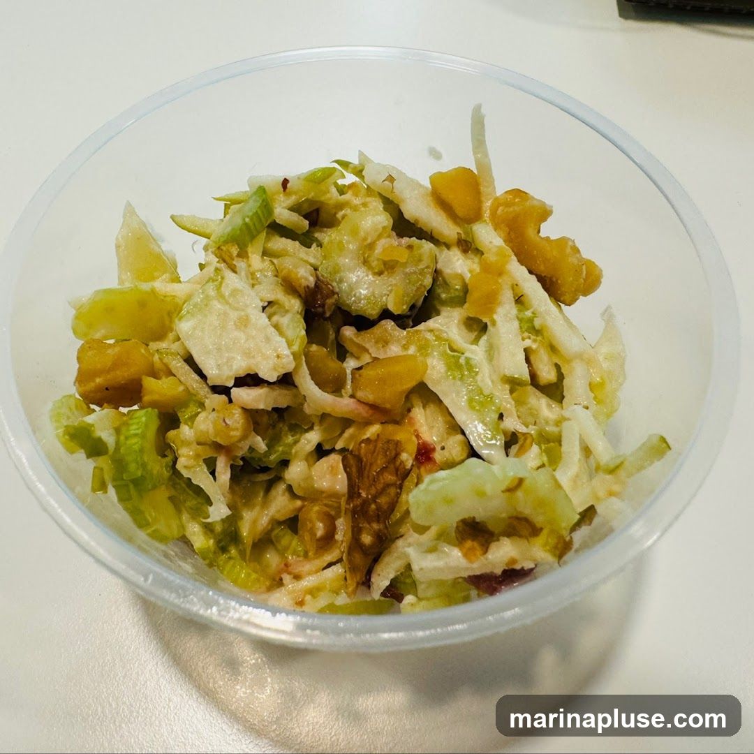 A close-up shot of a Waldorf salad in a clear plastic container. The salad appears to be made with chopped apples, celery, walnuts, and possibly raisins or dried cranberries, all coated in a creamy dressing.