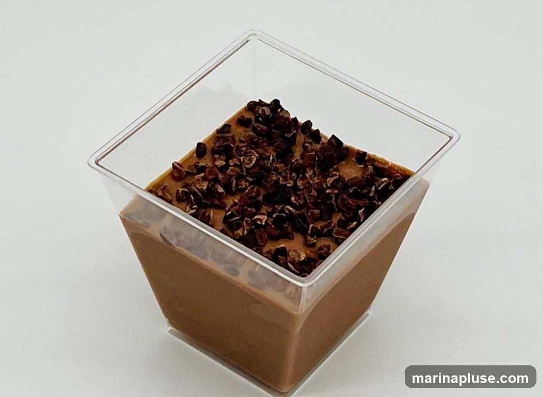 A close-up shot of a chocolate mousse dessert in a clear square plastic cup. The mousse is a smooth, rich brown color and is topped with a generous sprinkling of crushed cocoa nibs. The dessert is presented against a plain white background, highlighting its texture and color.