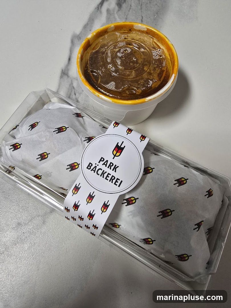 A clear plastic container holds a wrapped food item, possibly a pastry or sandwich, tied with a ribbon. The ribbon and the wrapping paper inside the container feature a repeating pattern of a stylized tulip-like graphic in red, yellow, and black. A circular sticker on the ribbon reads "PARK BÄCKEREI" with a similar graphic above the text. A white cup with a clear plastic lid contains a dark liquid, likely a beverage.