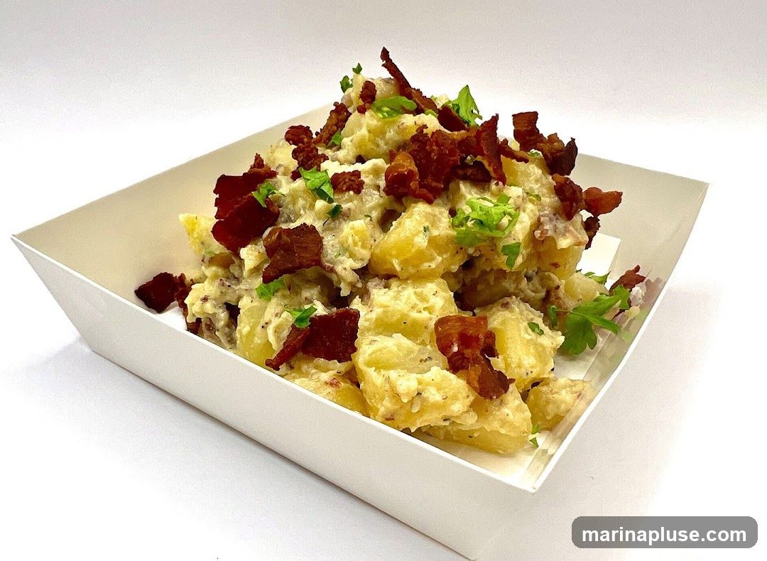 A white paper tray filled with potato salad. The salad is made with chunks of potato, a creamy dressing, and topped with crumbled bacon and chopped parsley.