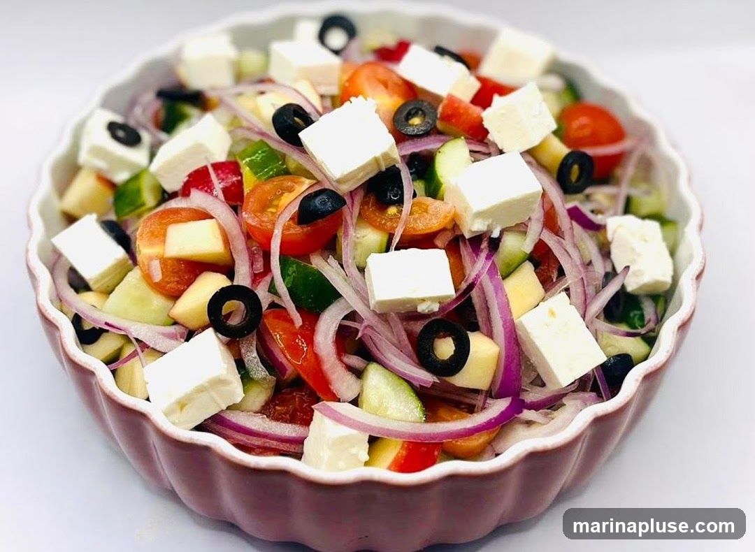 A close-up shot of a Greek salad in a decorative bowl. The salad is made with diced tomatoes, cucumbers, red onions, black olives, and cubes of feta cheese.