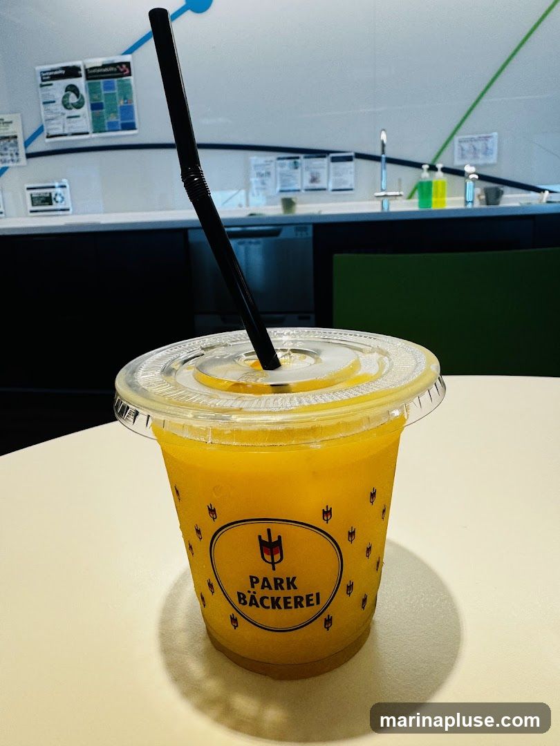 A close-up of a clear plastic cup filled with an orange beverage, likely a smoothie or juice, with a black straw. The cup has a repeating pattern of small tulip-like shapes and a central oval logo that reads "PARK BÄCKEREI" with a stylized graphic above it. The drink is on a white table, and the background shows a counter with a sink, soap dispensers, and some posters on the wall.