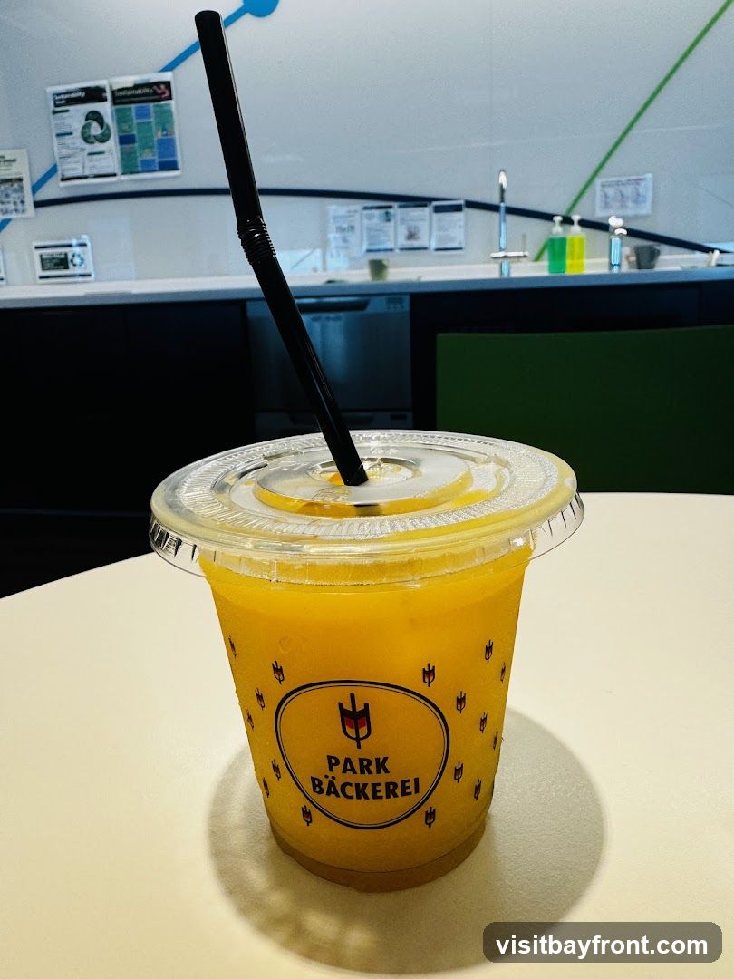 A close-up of a clear plastic cup filled with an orange beverage, likely a smoothie or juice, with a black straw. The cup has a repeating pattern of small tulip-like shapes and a central oval logo that reads "PARK BÄCKEREI" with a stylized graphic above it. The drink is on a white table, and the background shows a counter with a sink, soap dispensers, and some posters on the wall.