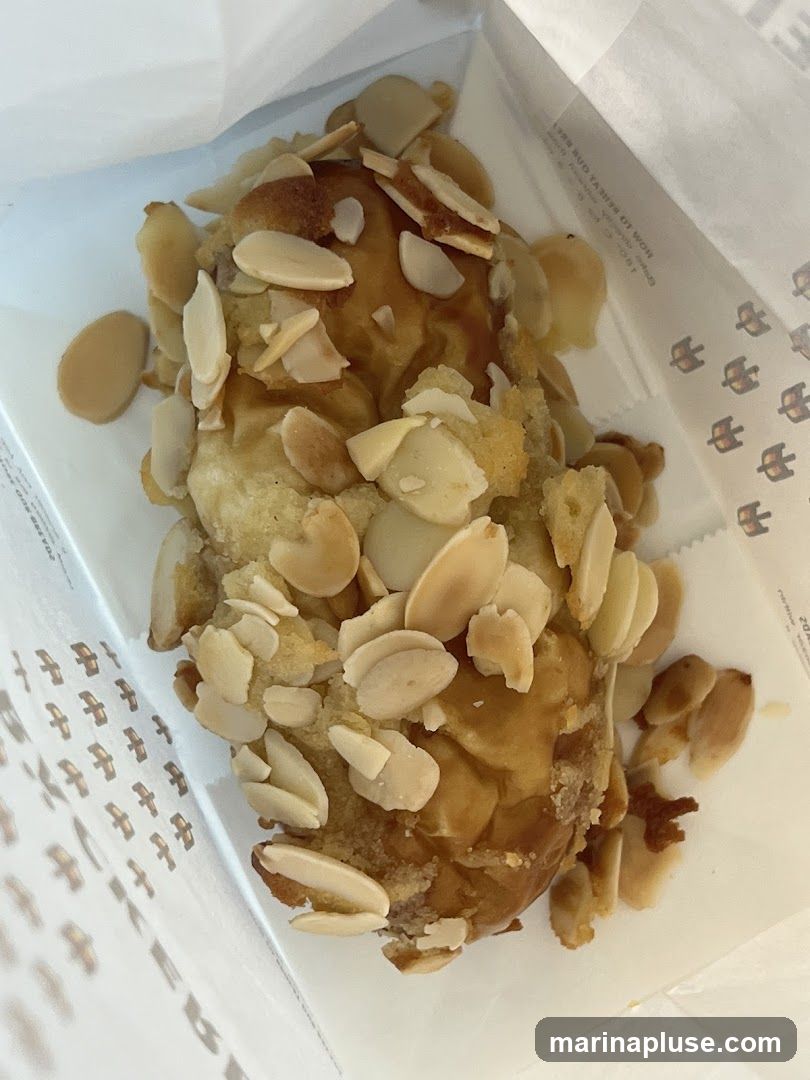A close-up shot of an almond croissant inside a white paper bag. The croissant is topped with sliced almonds and appears to have a glaze. The bag has a repeating pattern of small bird-like icons.