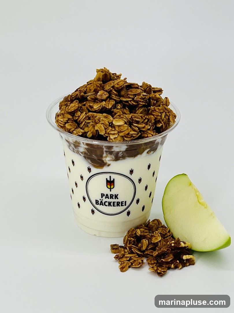 A clear plastic cup filled with yogurt, a layer of chocolate or pudding, and topped with a generous portion of granola. A slice of green apple and a small pile of granola are placed next to the cup. The cup has a logo that reads "PARK BÄCKEREI" with a stylized wheat stalk and German flag.