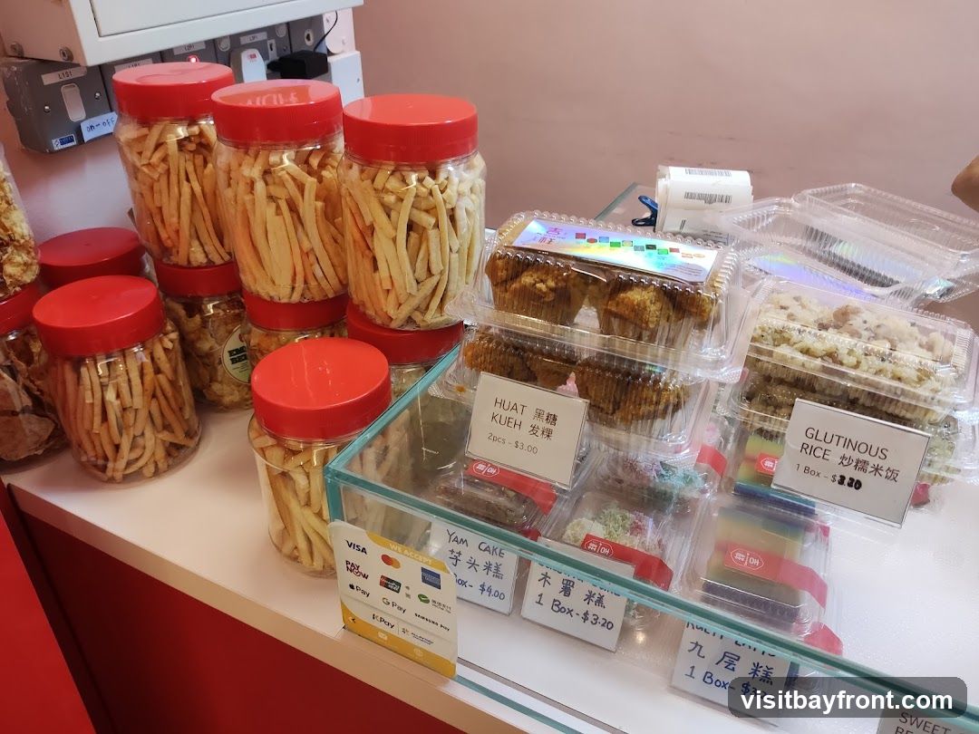 A display of various snacks in plastic containers and jars. In the foreground, there are clear plastic containers with layered cakes and muffins. Behind these, several large jars filled with thin, stick-like snacks are arranged. A price list is visible at the bottom left, indicating accepted payment methods and prices for items like Yam Cake, Huat Kueh, Glutinous Rice, and other cakes.