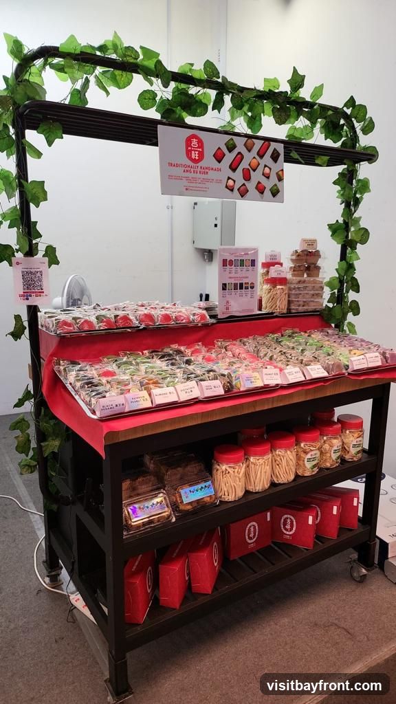 A display stand filled with various sweets and snacks. The stand is decorated with artificial green vines. On the top shelf, there are rows of individually packaged desserts. Below, more packaged desserts are displayed, with labels indicating flavors such as 'Black Sesame', 'Peanut', and 'Almond'. The bottom shelf holds red gift boxes and jars of snacks. A sign at the top advertises 'Traditionally Handmade Ang Ku Kueh' with images of the products.