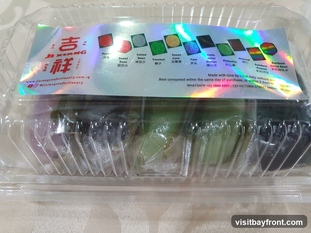 A clear plastic container filled with assorted pastries. The lid has a holographic label with the brand name "JI XIANG" and various flavors depicted with small images and text. Flavors include Peanut, Salted Bean, Sweet Bean, Coconut, Sweet Corn, Yam, 100% MSW Durian, Pistachio, Almond, Rainbow Peanut, and Rainbow Sweet Bean. The label also provides contact information and consumption advice.