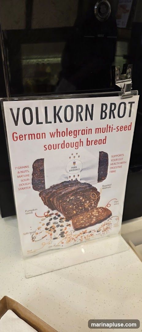 A sign advertising Vollkorn Brot, which is described as German wholegrain multi-seed sourdough bread. The sign features an image of the sliced bread and lists its ingredients, including 7 grains and nuts, mature sourdough starter, pumpkin seeds, split lentils, red quinoa, oat flakes, roasted walnuts, and flax seed. It also states that the bread supports gut health with digestive fibre.