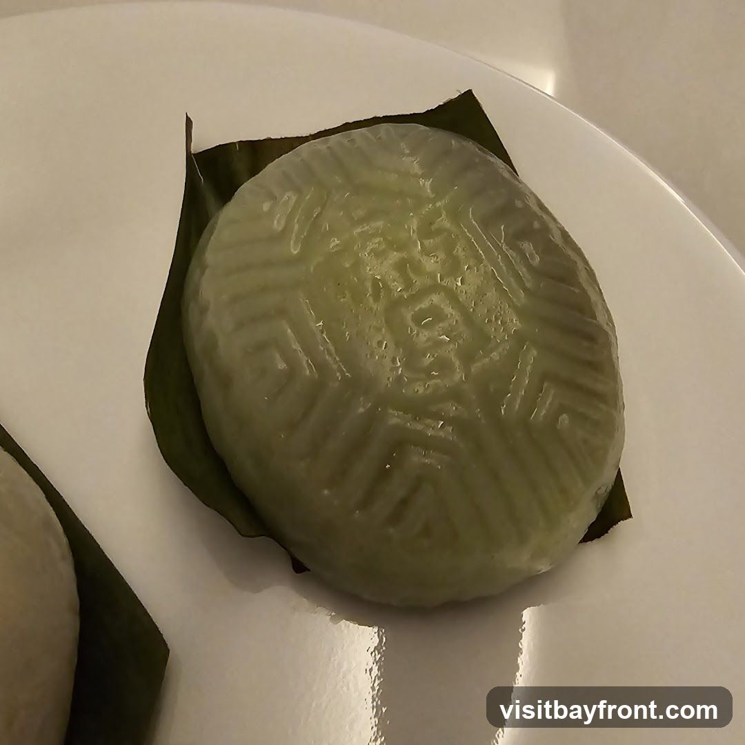 A close-up shot of a green, oval-shaped dessert resting on a dark green banana leaf. The dessert has a patterned surface with geometric designs. It is presented on a white plate. Another similar dessert is partially visible to the left.