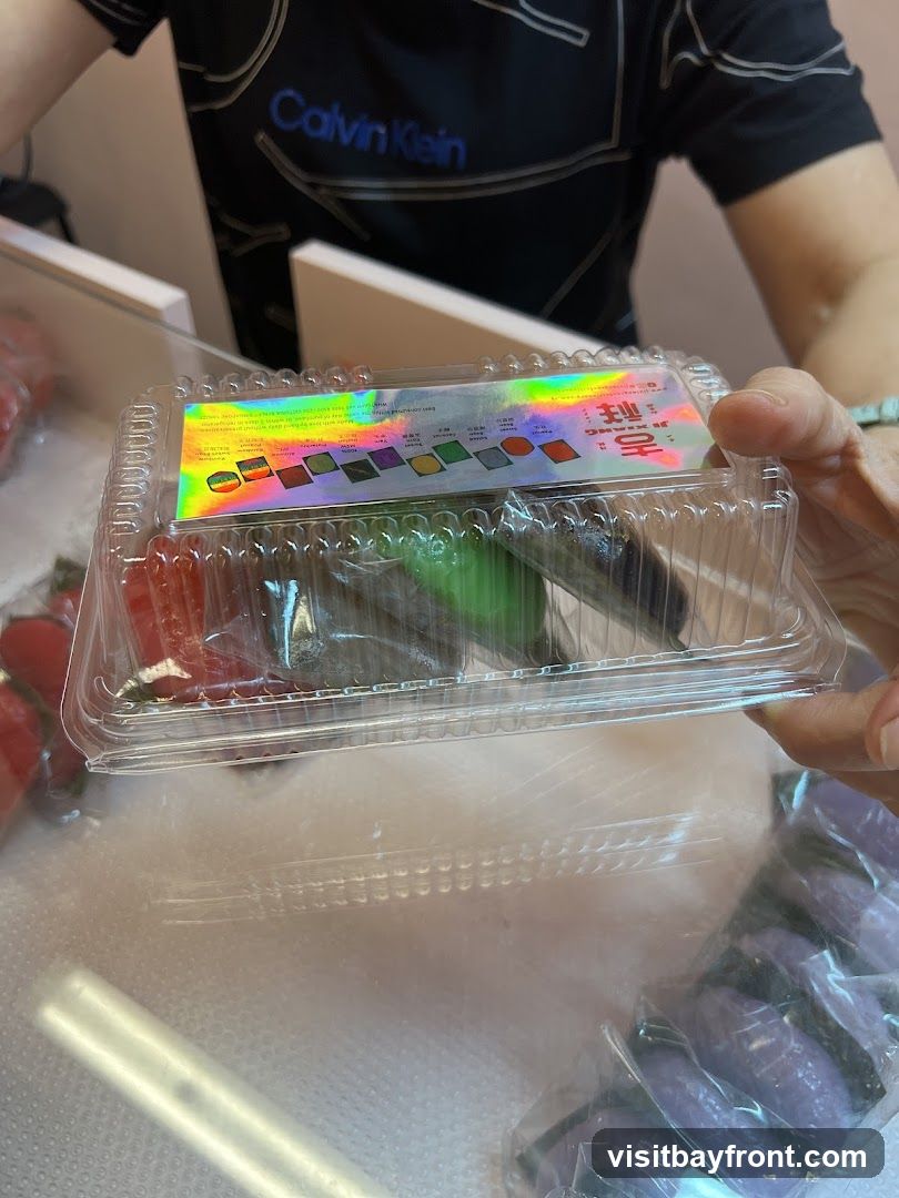 A clear plastic box containing various colorful desserts, possibly mochi or macarons. A holographic label on the lid displays the brand name "Ji Xiang" and images of different dessert flavors. In the background, a person wearing a Calvin Klein shirt is visible.