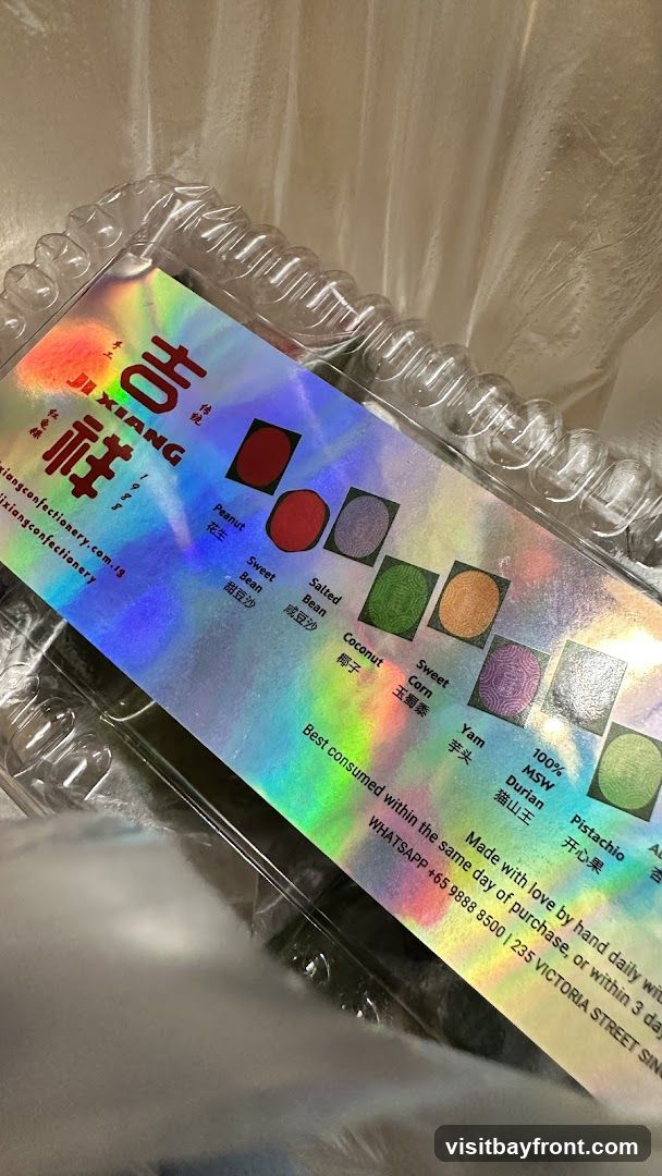A close-up of a clear plastic container holding confectionery items. The lid features a holographic label with the brand name "JI XIANG" in English and Chinese characters. The label displays small circular images representing different flavors, including Peanut, Sweet Bean, Salted Bean, Coconut, Sweet Corn, Yam, 100% MSW Durian, and Pistachio. Below the flavors, it states "Best consumed within the same day of purchase, or within 3 days" and provides contact information via WhatsApp and a physical address.