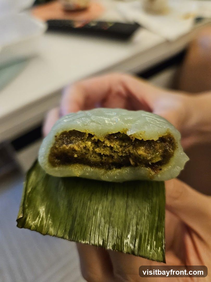 A close-up shot shows a person holding a sweet treat. The treat has a pale green, slightly translucent outer layer that has been bitten into, revealing a dark, granular filling. The treat is resting on a piece of green leaf, likely a banana leaf, which is also slightly shiny. The background is blurred, showing hints of a table and possibly a remote control.