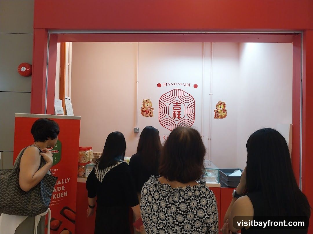 A group of people are standing in front of a stall with a red frame. The stall has a white background with some decorations. On the left side, there is a red banner with text that says "REALLY MADE KUEH CONFECTIONERY". There are also some jars of snacks visible. The people are looking at the items displayed in the stall.