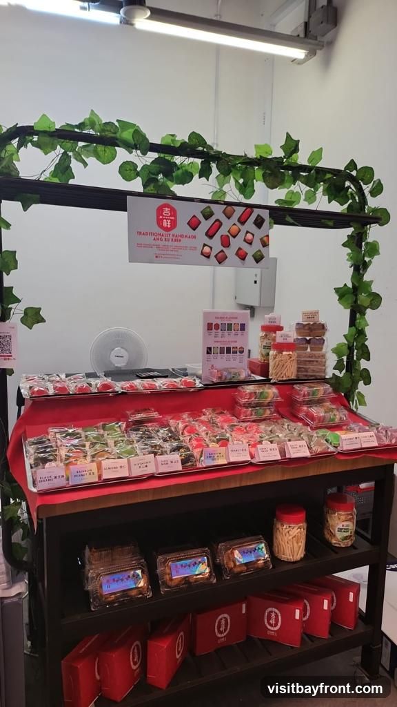 A display of various traditional handmade desserts, possibly cookies or pastries, arranged on shelves. The desserts are packaged in clear plastic containers and some are in red gift boxes. A sign above the display reads "TRADITIONALLY HANDMADE ANG KU KUEH" and shows images of the desserts. Artificial green vines are draped over a black frame above the display. A fan is visible in the background.