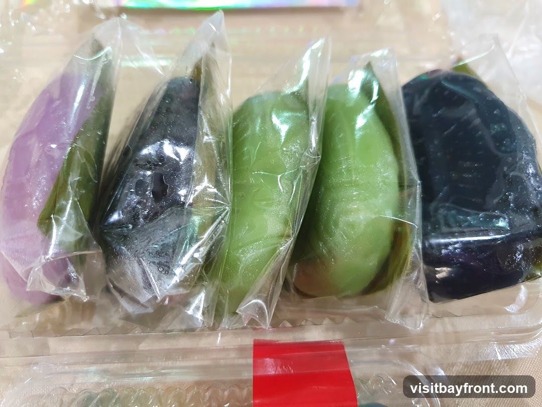 A close-up shot of five individually wrapped Japanese sweets in a clear plastic container. The sweets are cone-shaped and come in various colors: purple, dark green/black, and light green. They appear to be mochi or a similar confection.