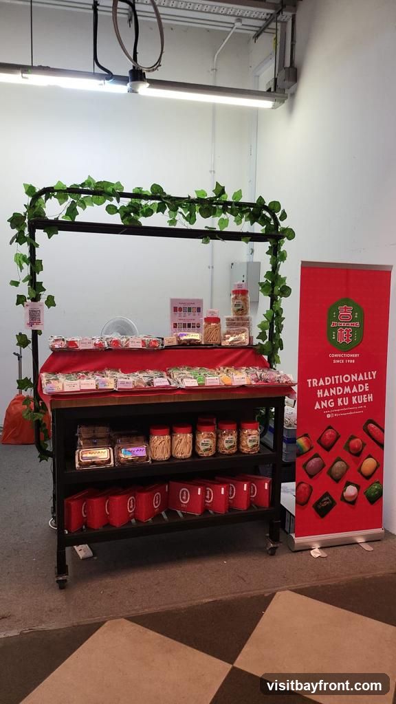 A display stand with various confectionery items, including Ang Ku Kueh, cookies, and other traditional snacks. A red banner on the right advertises "Traditionally Handmade Ang Ku Kueh" with images of the products. The stand is decorated with artificial green vines.