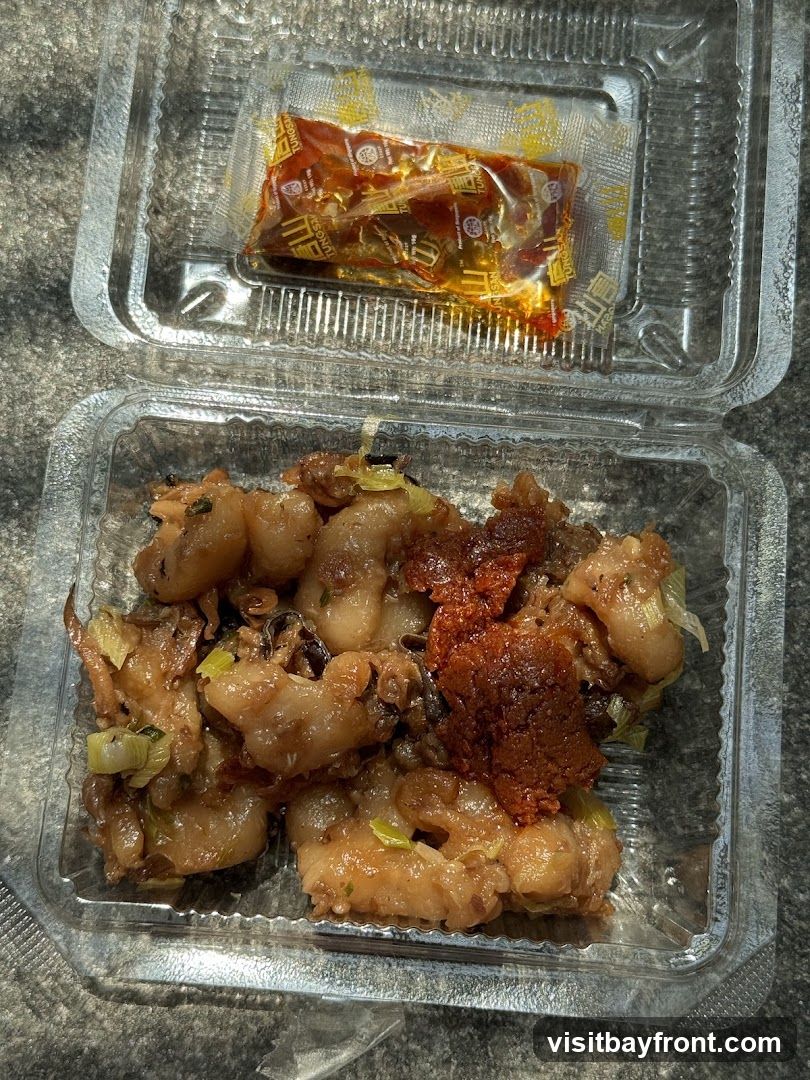 A clear plastic takeout container filled with a savory dish, possibly meat or seafood, mixed with green onions and a reddish paste. A small, sealed packet of sauce with yellow packaging and Chinese characters is placed on top of the food.