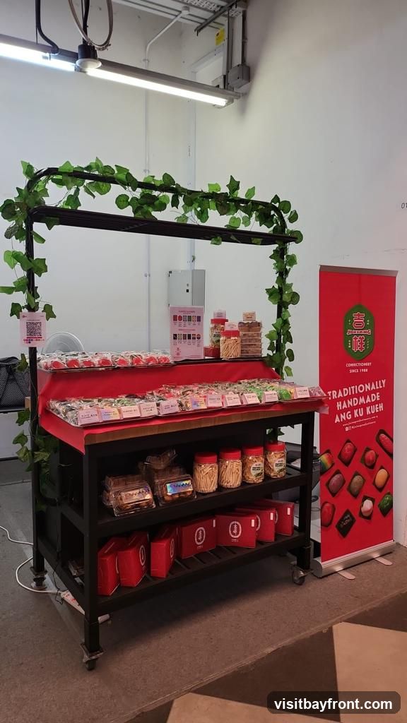 A display stand with various confectioneries, including Ang Ku Kueh, is set up indoors. The stand is decorated with artificial ivy. A red banner on the right advertises "Traditionally Handmade Ang Ku Kueh" and shows images of different flavors. There are also jars of snacks and red gift boxes on the shelves.