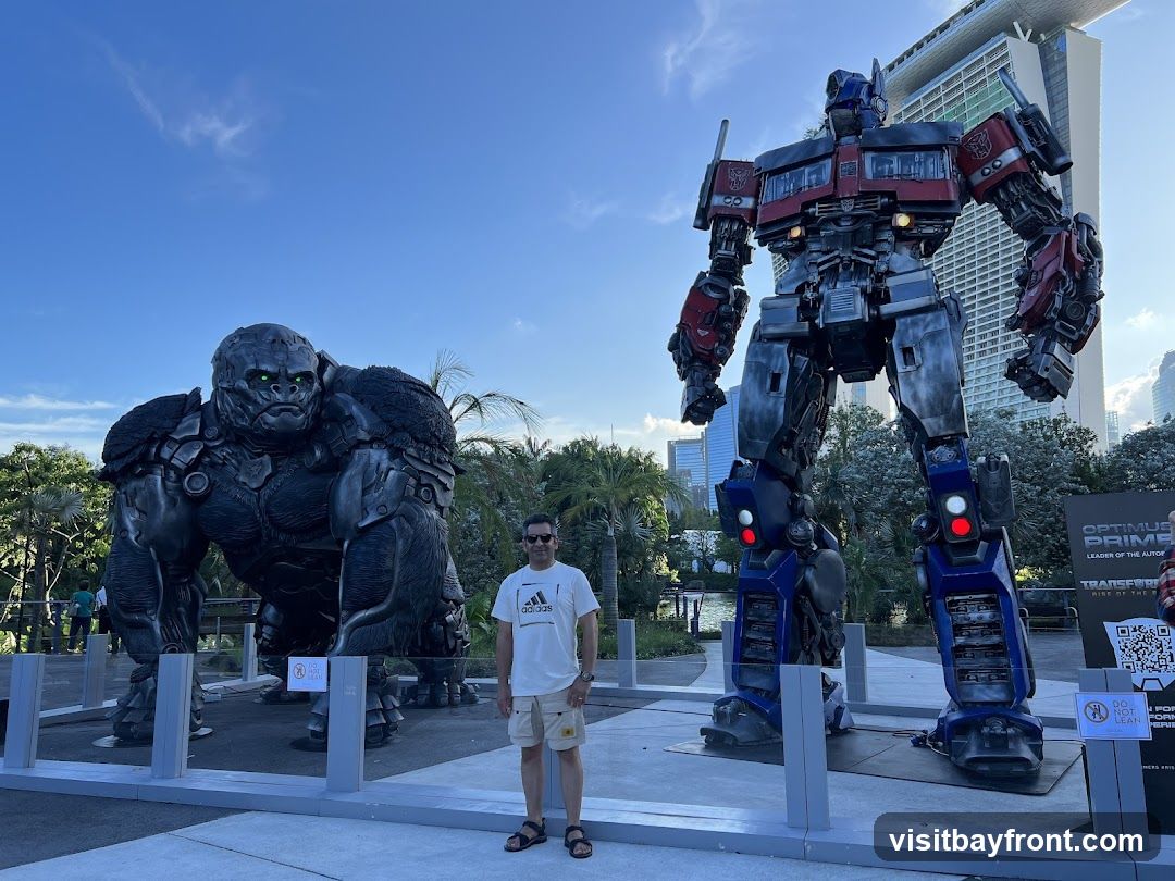 A man stands between two large statues: one of Optimus Prime from Transformers and the other of King Kong. Both statues are highly detailed and appear to be life-sized or larger. In the background, there are palm trees, modern buildings, and a bright blue sky with some clouds. A sign with a QR code and text related to "Optimus Prime" and "Transformers" is visible near the Optimus Prime statue.