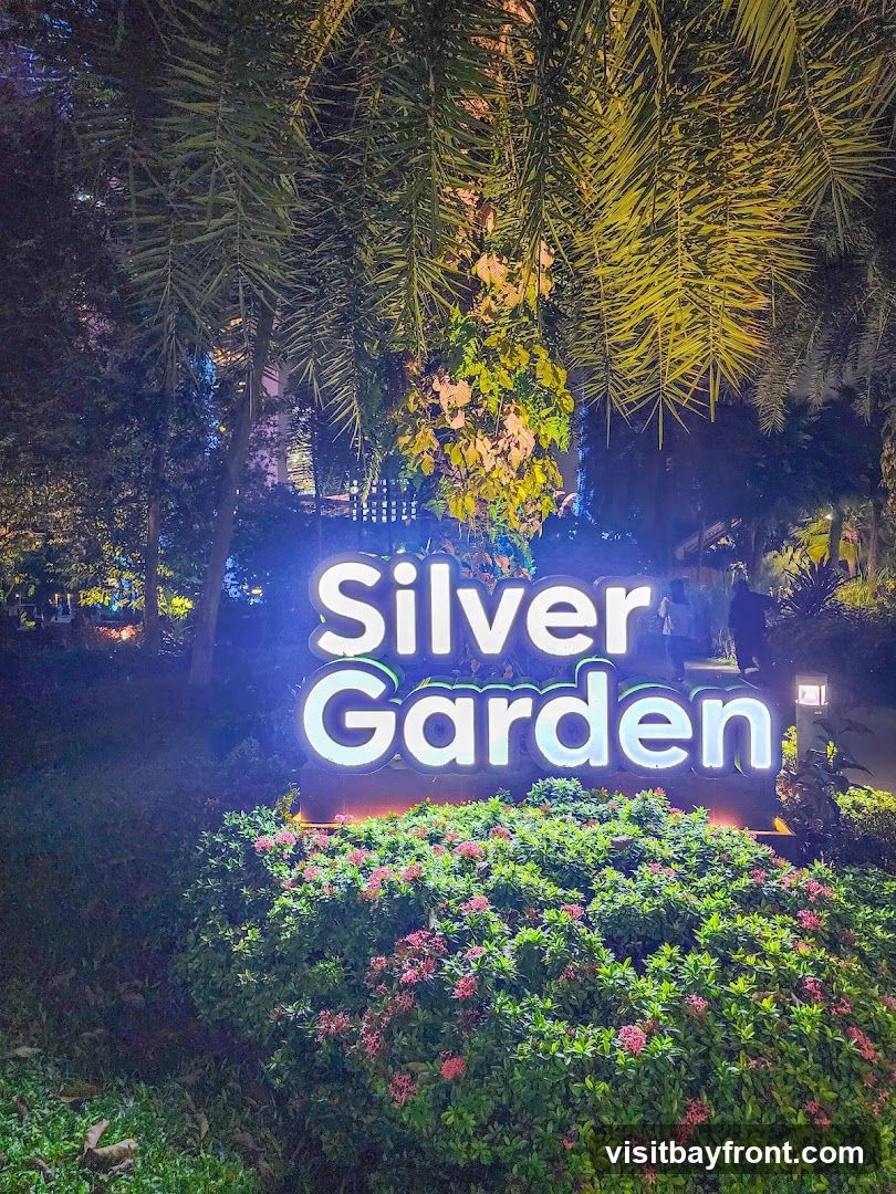 A large, illuminated sign that reads "Silver Garden" is set against a backdrop of lush greenery and palm fronds. The sign is lit with bright white light, making it stand out against the night sky. In front of the sign, there is a dense bed of flowering bushes with small pink blossoms. The overall scene suggests an outdoor garden or park area, possibly a venue or attraction.