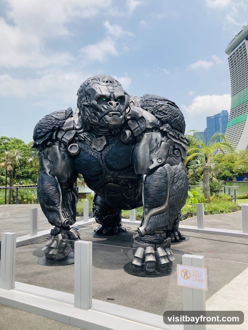 A large, metallic statue of a gorilla with glowing green eyes stands outdoors. The statue is intricately detailed, appearing to be made of mechanical parts. In the background, modern skyscrapers and lush greenery are visible under a partly cloudy sky. A small sign with a prohibition symbol and the text "DO NOT LEAN" is placed near the statue.