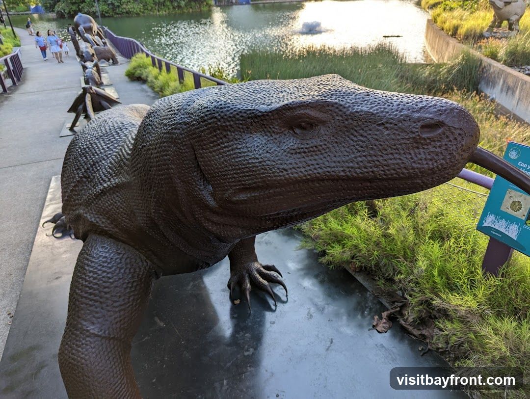 A large, dark brown sculpture of a Komodo dragon is positioned in the foreground, with its head and tongue extended. In the background, a body of water with a fountain is visible, along with lush green vegetation and other animal sculptures. Several people are walking along a path beside the water. A small informational sign with graphics is placed near the Komodo dragon sculpture.
