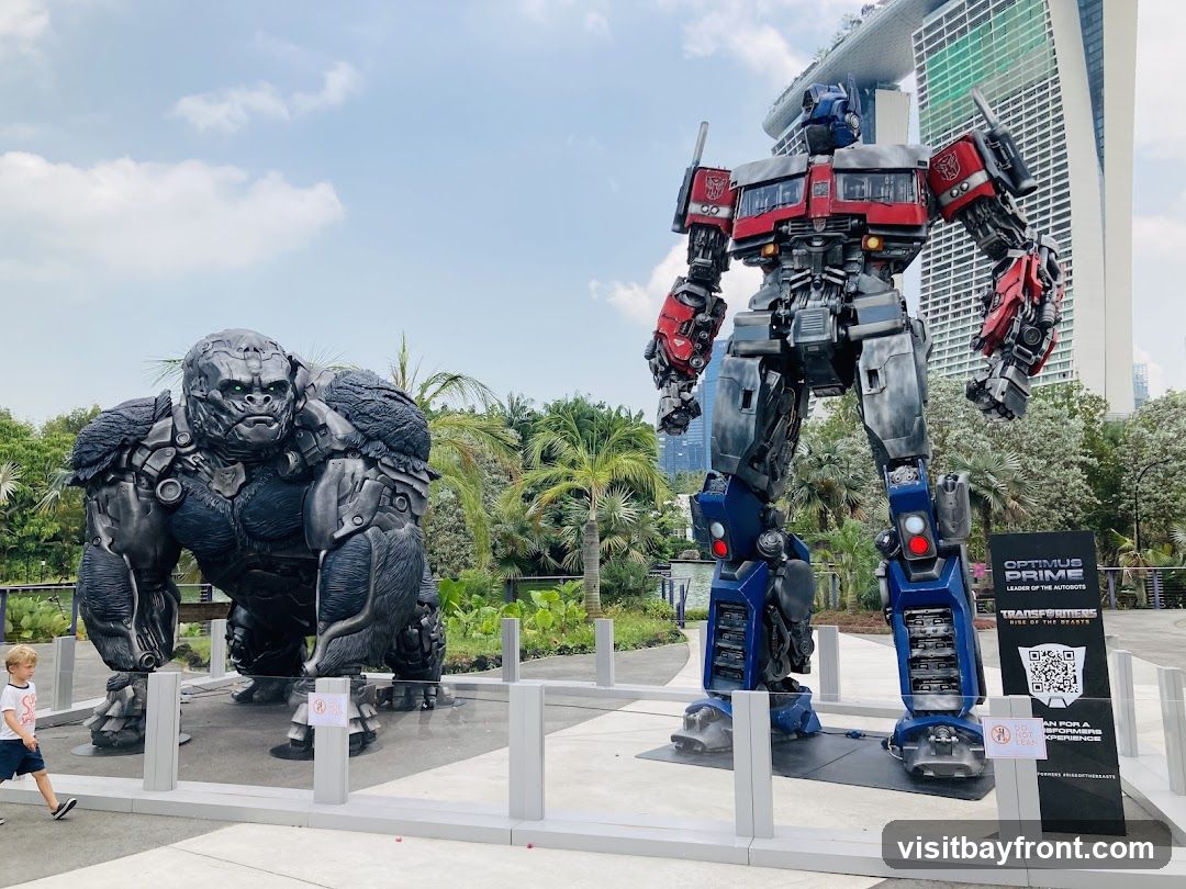 A large statue of Optimus Prime stands next to a statue of a robotic gorilla. In the background, a modern skyscraper and lush greenery are visible. A young boy walks past the gorilla statue. A sign near Optimus Prime reads "OPTIMUS PRIME LEADER OF THE AUTOBOTS TRANSFORMERS RISE OF THE BEASTS" and includes a QR code.
