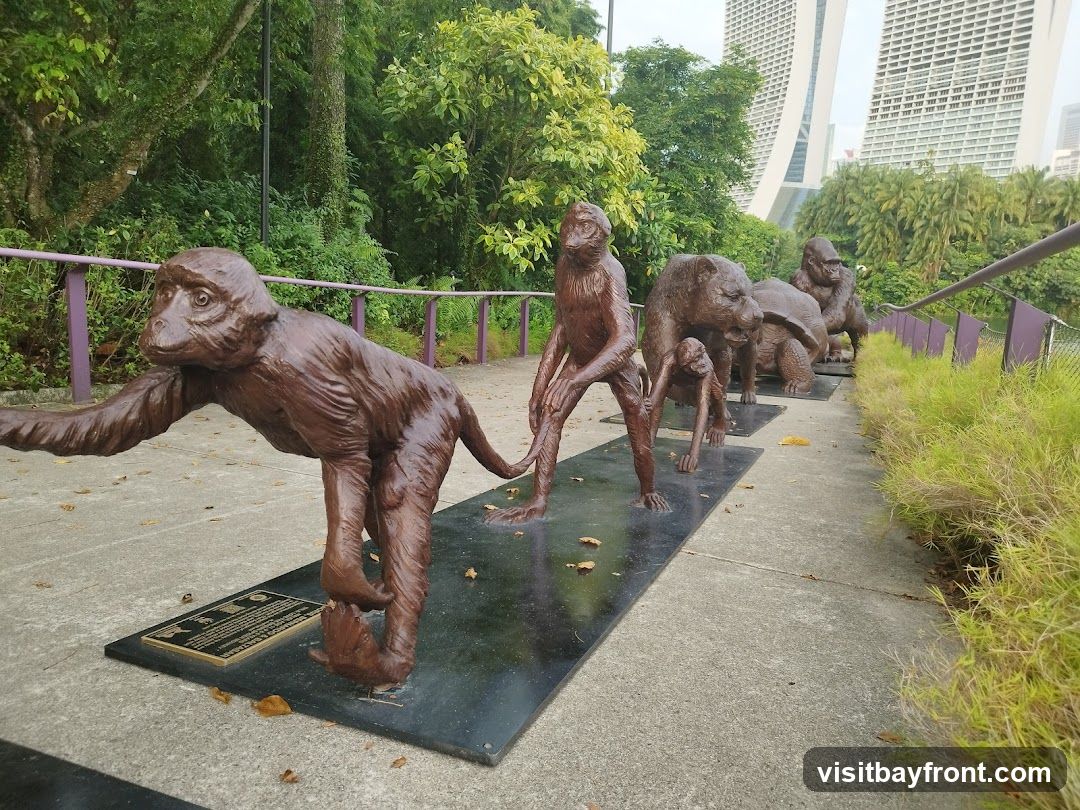 A series of bronze animal sculptures, including monkeys, a bear, a turtle, and a gorilla, are displayed in a line on a paved walkway. The sculptures are placed on black rectangular bases. In the background, lush green foliage and modern skyscrapers are visible. A purple railing runs along the side of the walkway, and dry leaves are scattered on the ground.