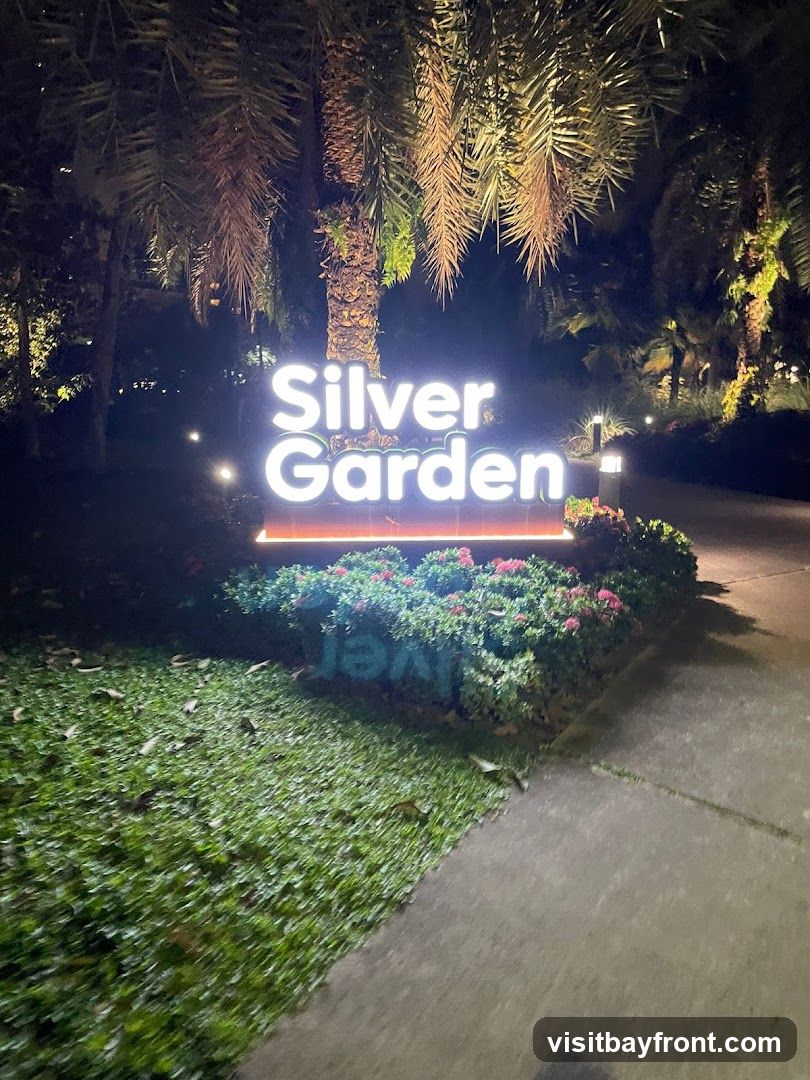A brightly lit sign that reads "Silver Garden" is set against a backdrop of palm trees and lush greenery at night. The sign is illuminated with white light, and a subtle orange glow emanates from its base. The surrounding area is softly lit by spotlights, highlighting the tropical foliage. A paved walkway is visible in the foreground, with a patch of grass to the left.