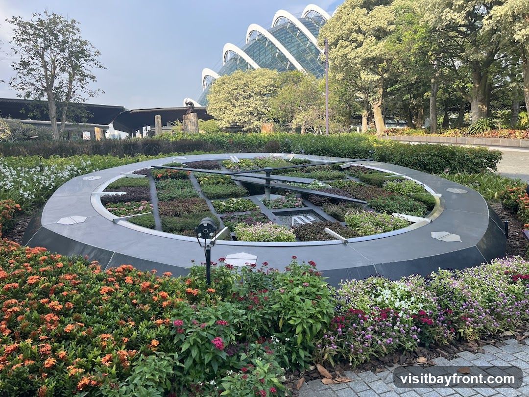 A large, circular floral clock is set amidst a garden with various flowers and lush greenery. In the background, a distinctive geodesic dome structure with a white, arched roof is visible, along with trees and other garden elements. The clock face is filled with different types of plants, and its hands indicate the time.