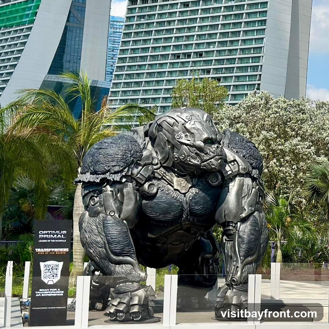 A large, metallic statue of Optimus Primal, a character from Transformers: Rise of the Beasts, is displayed outdoors. The statue resembles a gorilla with mechanical enhancements. In the background, modern buildings and lush greenery are visible. A sign next to the statue provides information about Optimus Primal and the movie, along with a QR code for an AR experience.