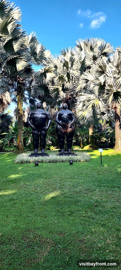 Two dark, stylized human figures stand on a grassy lawn surrounded by lush palm trees and tropical foliage. The sky above is a clear blue with a few wispy clouds. A small sign is visible to the right of the sculptures.