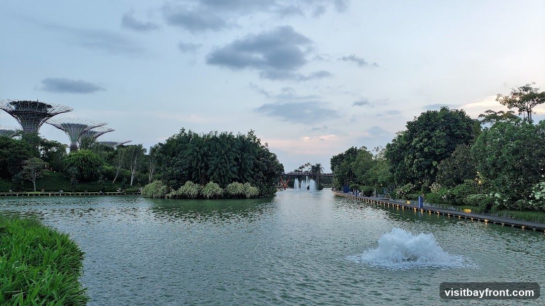 A scenic view of Gardens by the Bay in Singapore, featuring the iconic Supertree Grove in the distance, a tranquil body of water with a fountain, and lush greenery. The sky is overcast with soft clouds, suggesting it might be late afternoon or early evening.