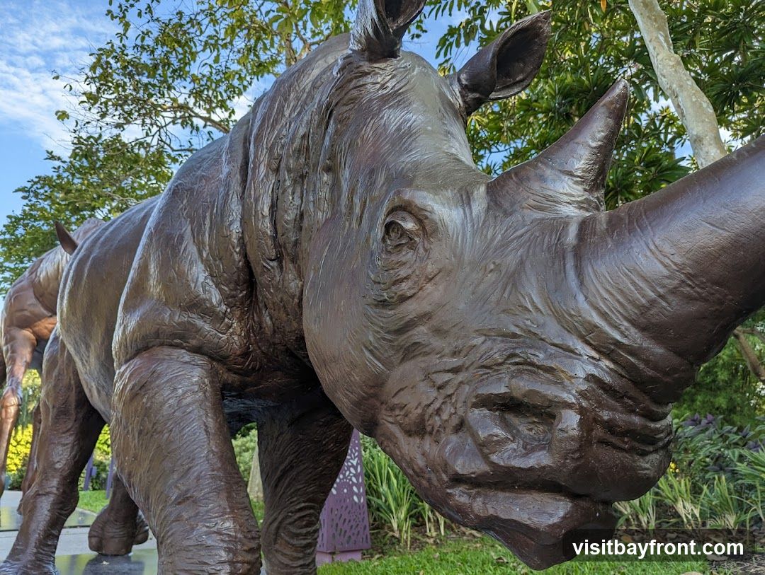 A close-up, low-angle shot of a large, dark brown rhinoceros sculpture. The sculpture is detailed, showing the texture of the rhino's skin, its horn, ears, and eye. It is positioned outdoors with lush green foliage and a blue sky with clouds visible in the background. Another sculpture, possibly of a horse, is partially visible behind the rhino.