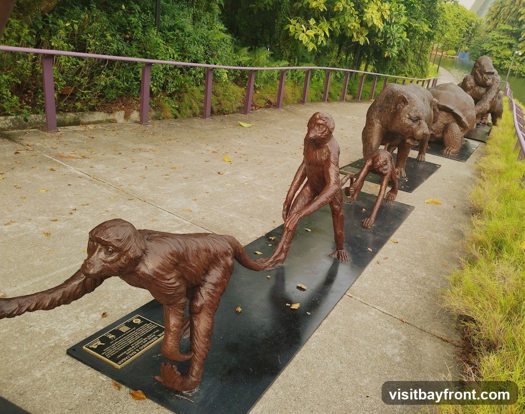 A line of bronze animal sculptures, including monkeys, a bear, a turtle, and a gorilla, are displayed on a paved walkway. The sculptures are placed on black rectangular bases. A purple railing runs along the left side of the walkway, with lush green foliage behind it. To the right, there is a body of water and more greenery.