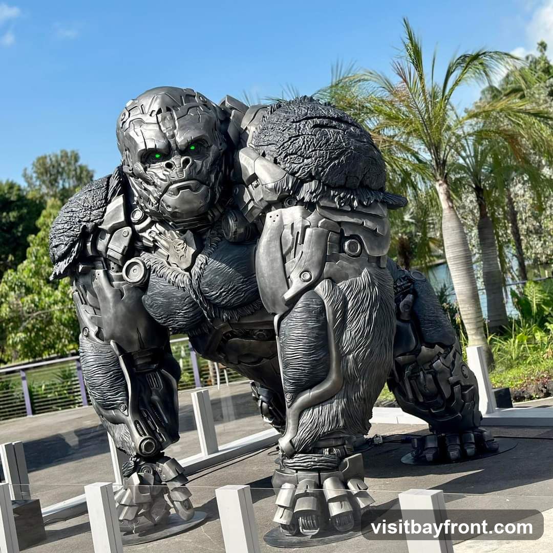 A large, metallic sculpture of a gorilla with glowing green eyes stands outdoors. The sculpture is detailed, with visible mechanical components and textured fur-like elements. Palm trees and lush greenery are visible in the background under a clear blue sky.