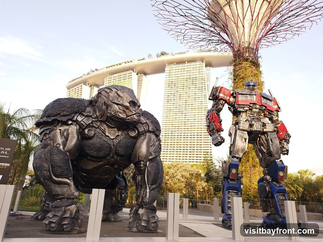 A large statue of Optimus Prime from Transformers stands next to a statue of a gorilla. In the background, the Marina Bay Sands hotel and a Supertree Grove from Gardens by the Bay are visible.