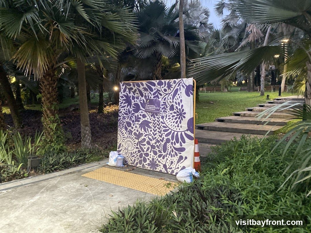 A large display board with a purple floral pattern stands in a park-like setting. The board has text on it that reads, "We are making improvements to the gardens." It is propped up by a yellow frame and secured with white bags at its base. An orange traffic cone is placed next to it. The background features lush greenery, palm trees, and a set of concrete stairs leading up a grassy slope.
