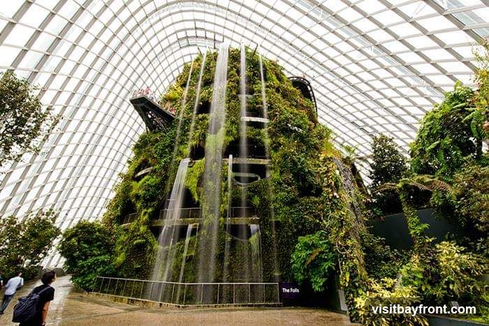 An indoor botanical garden with a large, multi-tiered structure covered in lush greenery and cascading waterfalls. A glass and metal geodesic dome roof covers the space. Several people are visible, including two walking in the foreground and a group on a walkway near the top of the structure. A sign reads "The Falls".