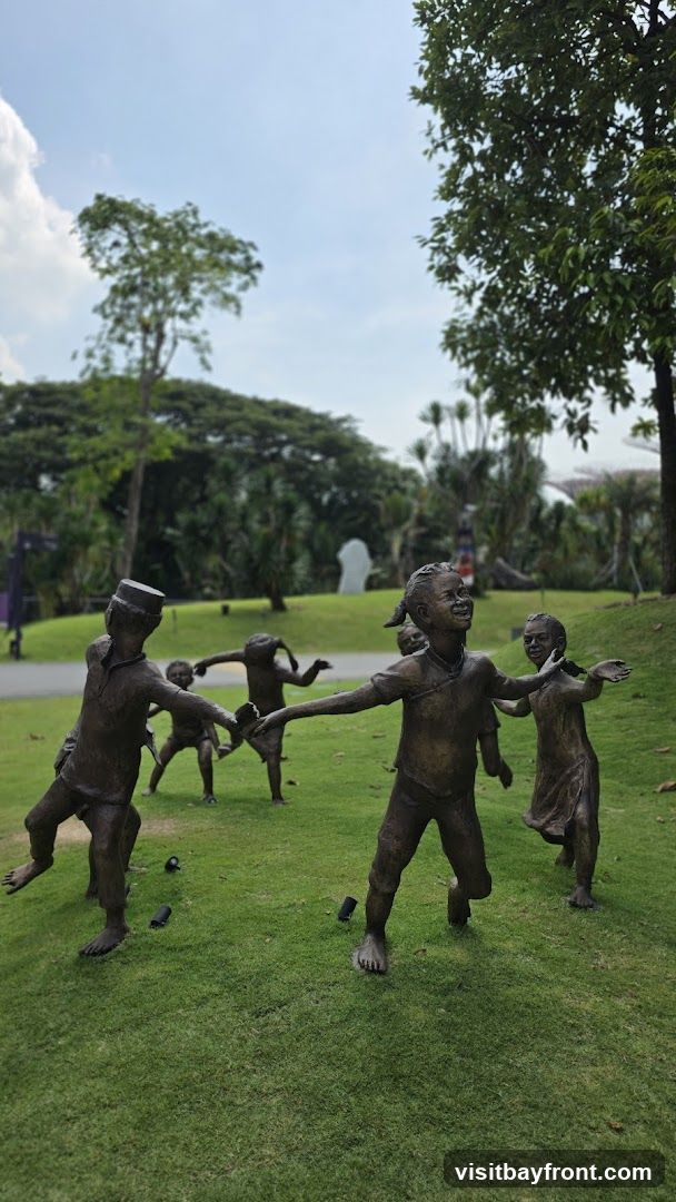A bronze sculpture depicting several children playing a game of ring around the rosy on a grassy lawn. The children are in various poses of movement and joy. In the background, there are trees, lush greenery, and a cloudy sky.