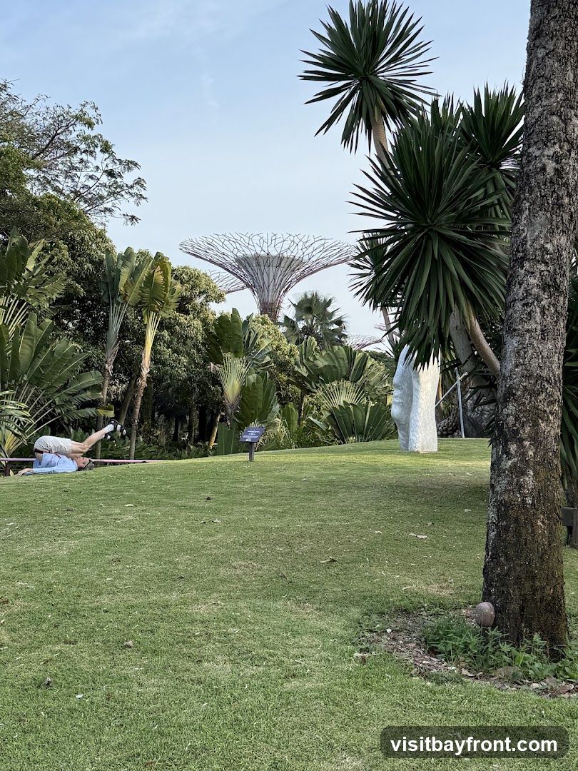A person is doing yoga on a tightrope in a grassy area with lush tropical plants and trees. In the background, the iconic Supertree Grove structures of Gardens by the Bay are visible under a clear sky.