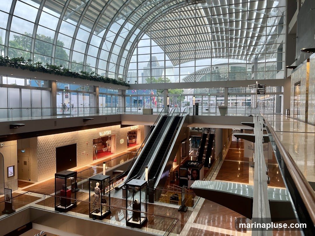 The image shows the interior of a spacious, modern shopping mall with a high, arched glass ceiling. Escalators are visible, connecting different levels. Several luxury brand stores, including Valentino and Chanel, are identifiable by their signage and displays. Mannequins are showcased in glass display cases. The mall features polished floors and contemporary architecture.